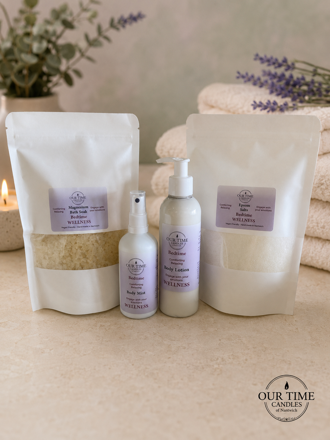 A set of natural skincare products from Our Time Candles of Nantwich, including two white stand-up pouches of bath soak and Epsom salts, a pump bottle of body lotion, and a spray bottle of body mist, arranged on a beige stone surface with a candle, folded towels, and lavender sprigs in the background.