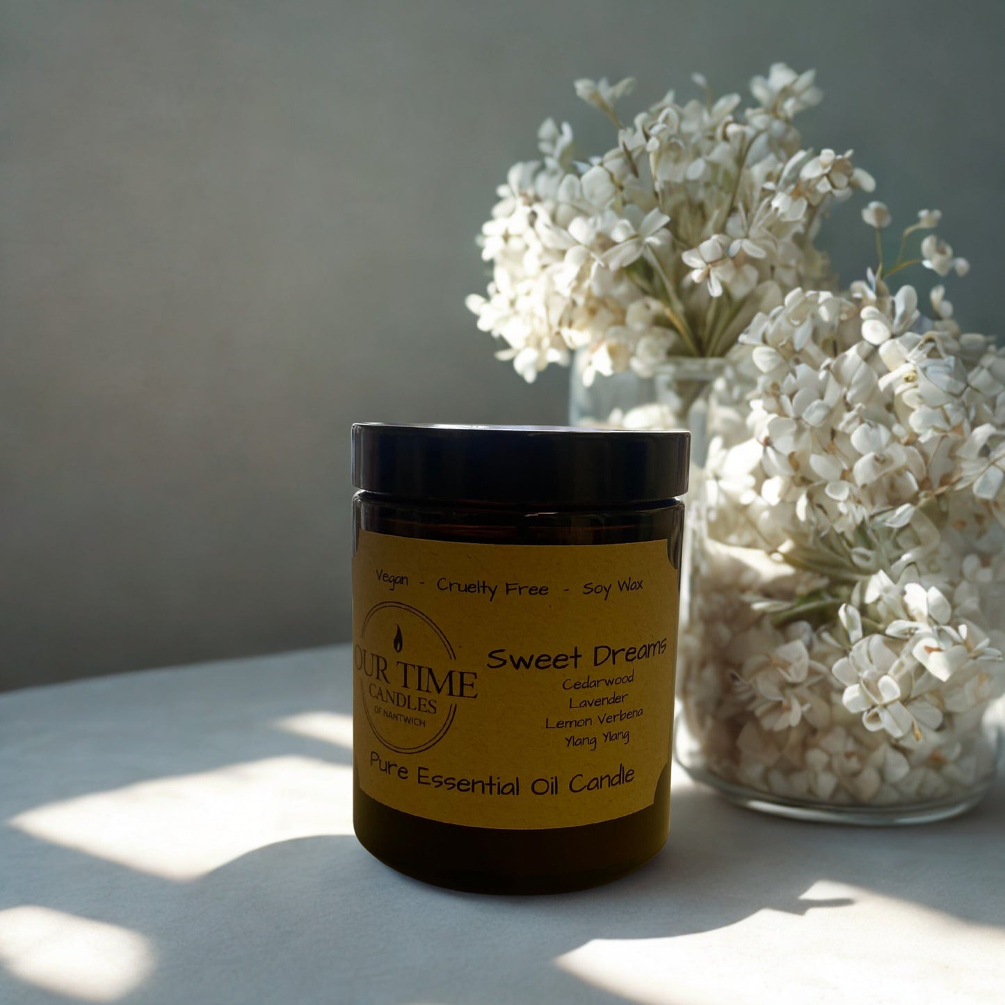 Candle labeled 'Sweet Dreams' with a vase of white flowers on a light surface.